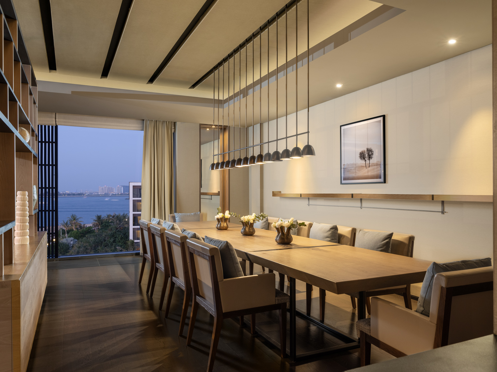 Modern dining room with a long wooden table, cushioned chairs, and minimalist pendant lighting. Floor-to-ceiling windows offer a scenic ocean view at sunset, while soft neutral tones and elegant décor create a warm, sophisticated atmosphere.